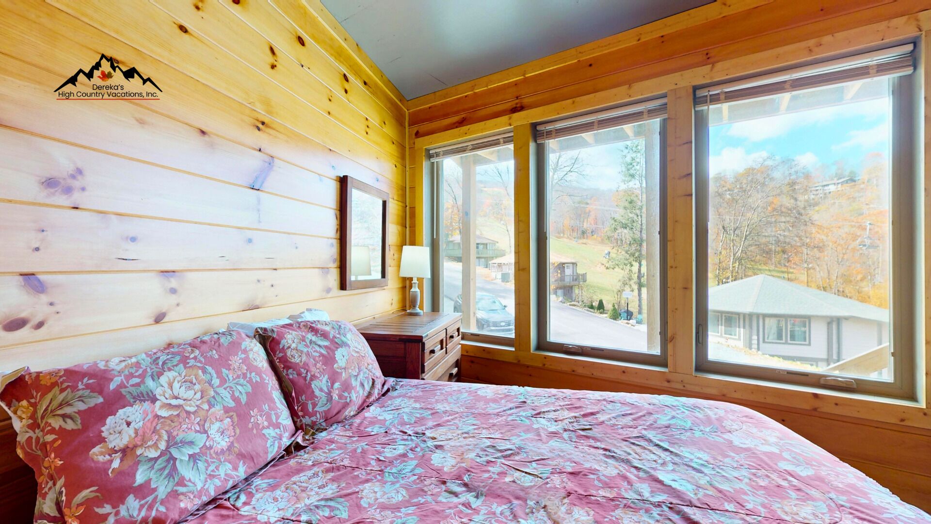 Ein Zimmer in einer Holzhütte mit einem Bett, einem Nachttisch und großen Fenstern mit Blick nach draußen.