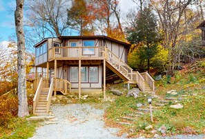 A wooden cabin with a balcony and stairs, surrounded by trees and a gravel path.