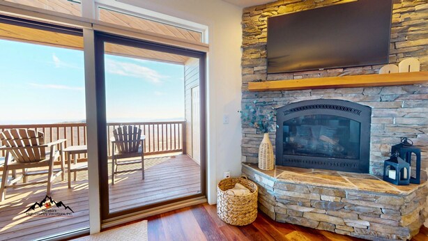 A room with a stone fireplace, a flat-screen TV mounted above it, and a balcony with wooden deck and chairs. - Reserve 36C • Top of the World • Hot Tub (Sugar Mountain)
