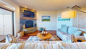 A living room with a stone fireplace, a flat-screen TV mounted above it, a sofa with patterned cushions, a round wooden coffee table, and a large window with a view of the outdoors. - Reserve 36C • Top of the World • Hot Tub (Sugar Mountain)