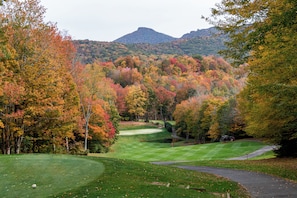 A golf course with a pond, surrounded by autumn-colored trees and hills in the background.