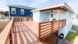 Una terraza de madera con barandilla, un área de estar al aire libre techada y un edificio azul claro con balcón.