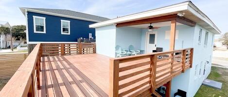 Une terrasse en bois avec une rambarde, un espace extérieur aménagé et couvert, et un bâtiment bleu clair doté d’un balcon.