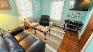 A living room with a black leather sofa, a wicker chair, a black armchair, a small round table, and a TV mounted on the wall. - Gallows Hill Guest House, Charming 2BR Apartment on Gallows Hill in Salem, MA (Salem)