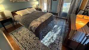 A bedroom with a large bed, a nightstand, a lamp, a window with curtains, and a wall-mounted artwork. - Gallows Hill Guest House, Charming 2BR Apartment on Gallows Hill in Salem, MA (Salem)