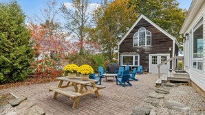 A wooden house with a large window, a patio with a picnic table and blue chairs, and a garden with trees and flowers.