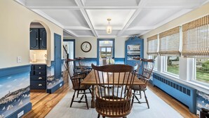 A dining area with a wooden table and chairs, a clock on the wall, and a view of the outdoors through large windows.