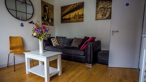 Un salón con una mesa blanca, un sofá negro y un jarrón con flores. Hay cuadros en la pared y una puerta con un letrero.