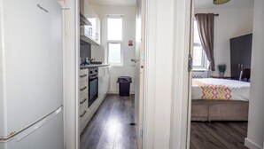 Un pasillo estrecho que conduce a una cocina con refrigerador, horno y alacenas, y un dormitorio con cama y ventana.