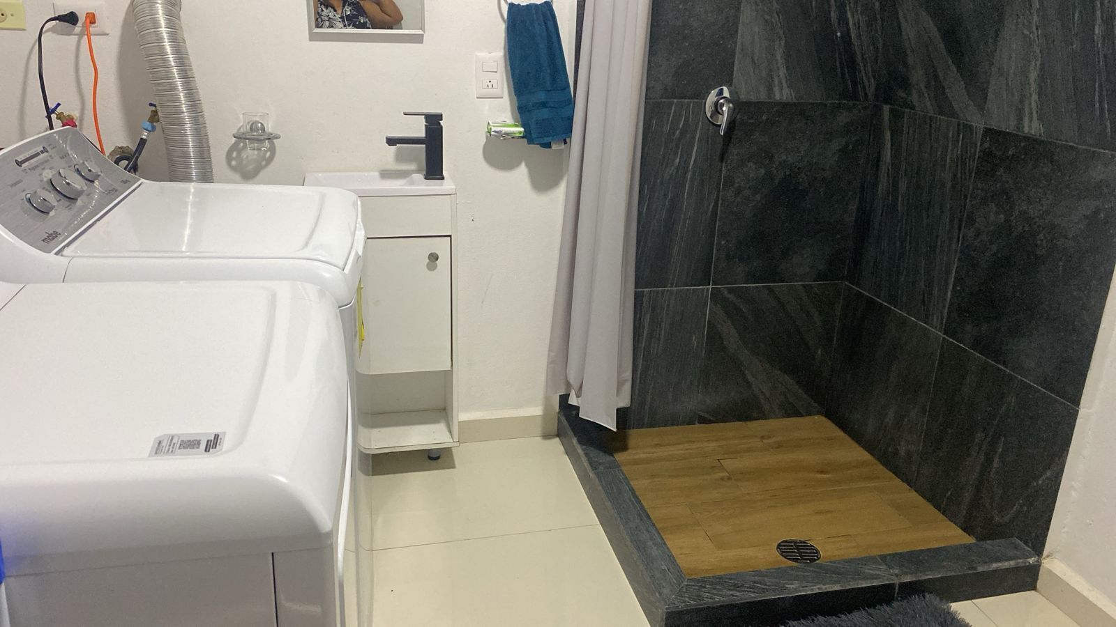 Een badkamer met een witte wasmachine, een klein kastje, een douche met donkere tegels en een houten vloer.