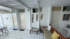A clean, white interior with a tiled floor, a wooden bench with cushions, a small table, and a potted plant. - Cozy Room w/ Private Bath + Shared Kitchen 5 Min to Beach -Surf Hostel Vibe (San Miguel)