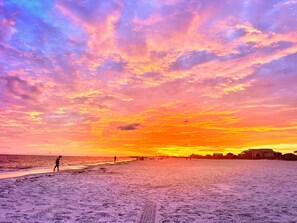Ein Strand bei Sonnenuntergang, eine Person geht spazieren, und Gebäude sind in der Ferne sichtbar.
