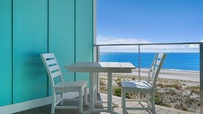 Ein Balkon mit Tisch und zwei Stühlen, mit Blick auf einen Strand und das dahinterliegende Meer.