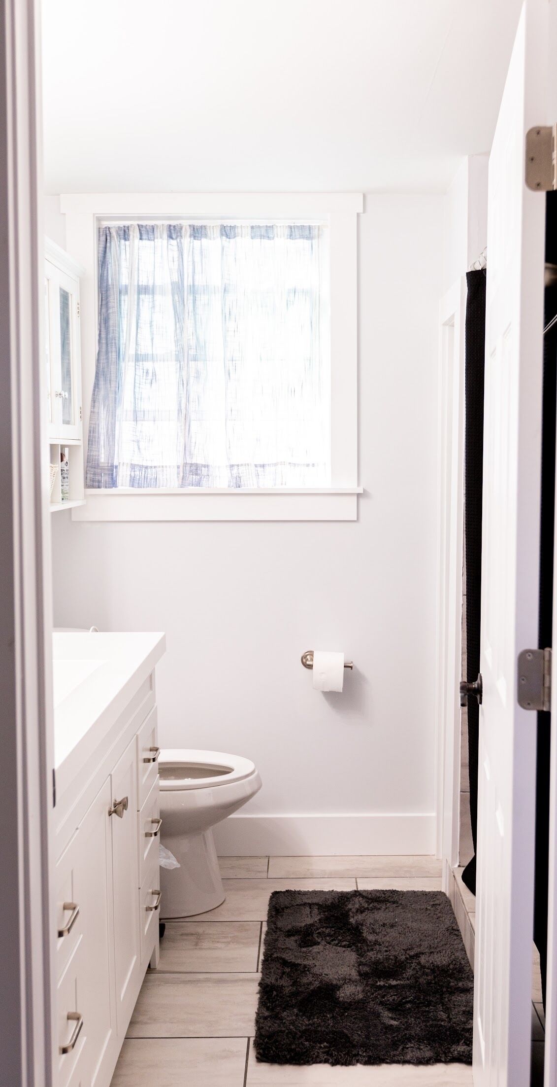A small, white bathroom with a toilet, a white sink, and a window with a blue curtain.
