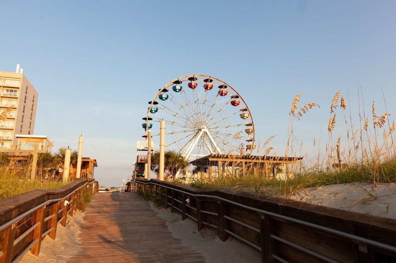 Een vogelvluchtperspectief van een looppad dat leidt naar een groot reuzenrad, met een gebouw en strandbeplanting op de achtergrond.