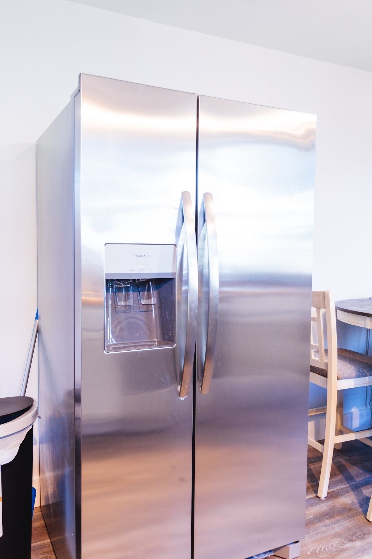 A stainless steel refrigerator with a water dispenser, located in a room with a wooden dining table and chairs.