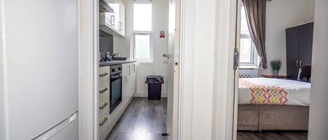 A narrow hallway leading to a kitchen with a refrigerator, oven, and cabinets, and a bedroom with a bed and a window.