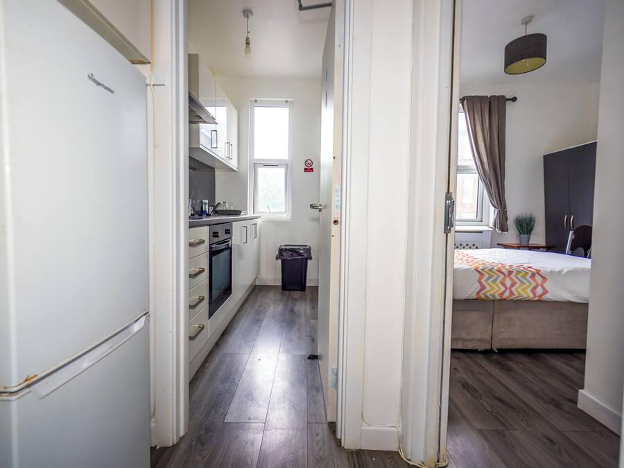 A narrow hallway leading to a kitchen with a refrigerator, oven, and cabinets, and a bedroom with a bed and a window.