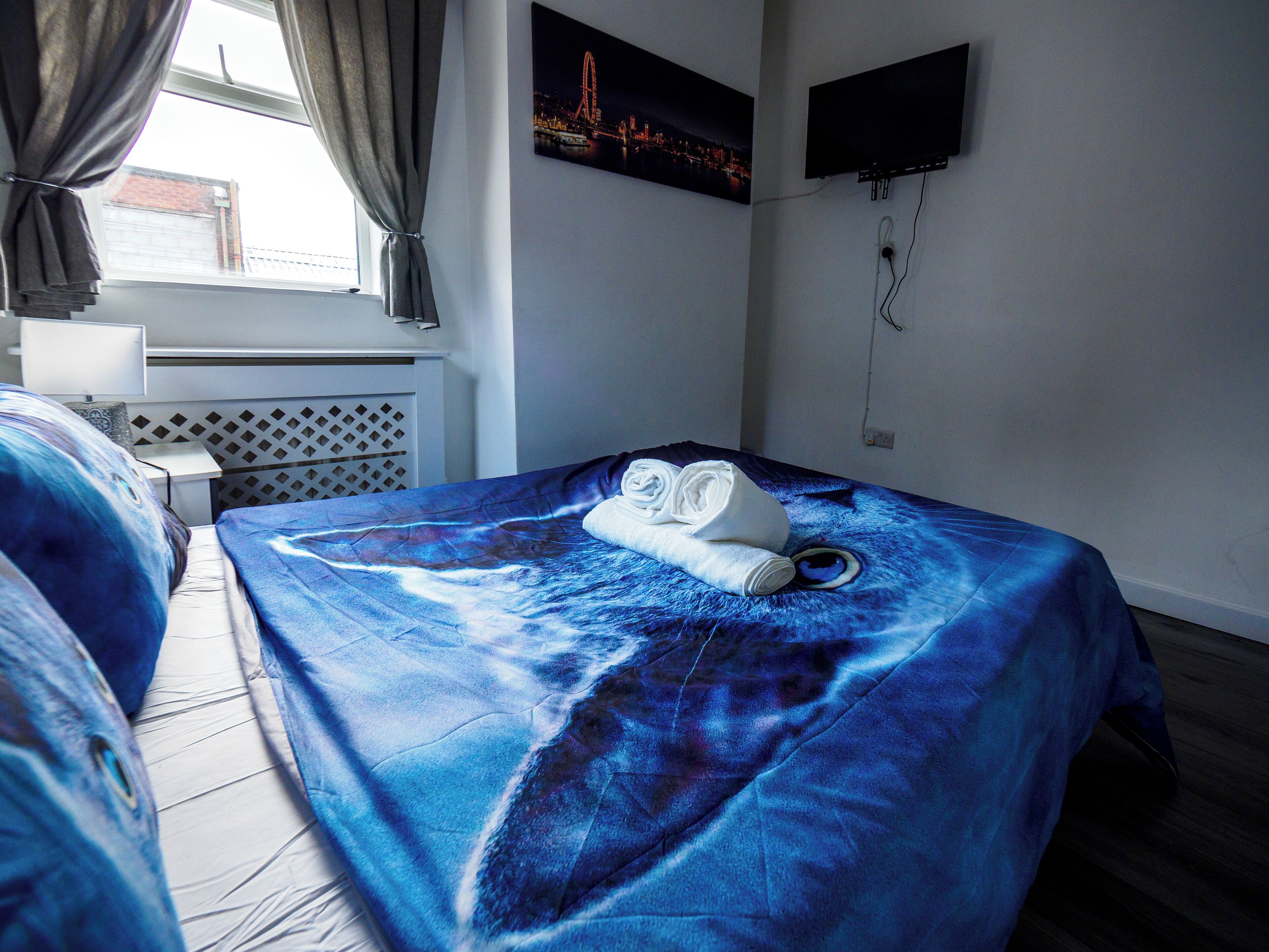 A hotel room with a bed covered in a blue ocean-themed bedspread, a neatly folded towel on the bed, a flat-screen TV mounted on the wall, and a window with curtains.