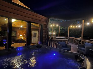 A hot tub with a wooden deck, a covered seating area, and string lights.