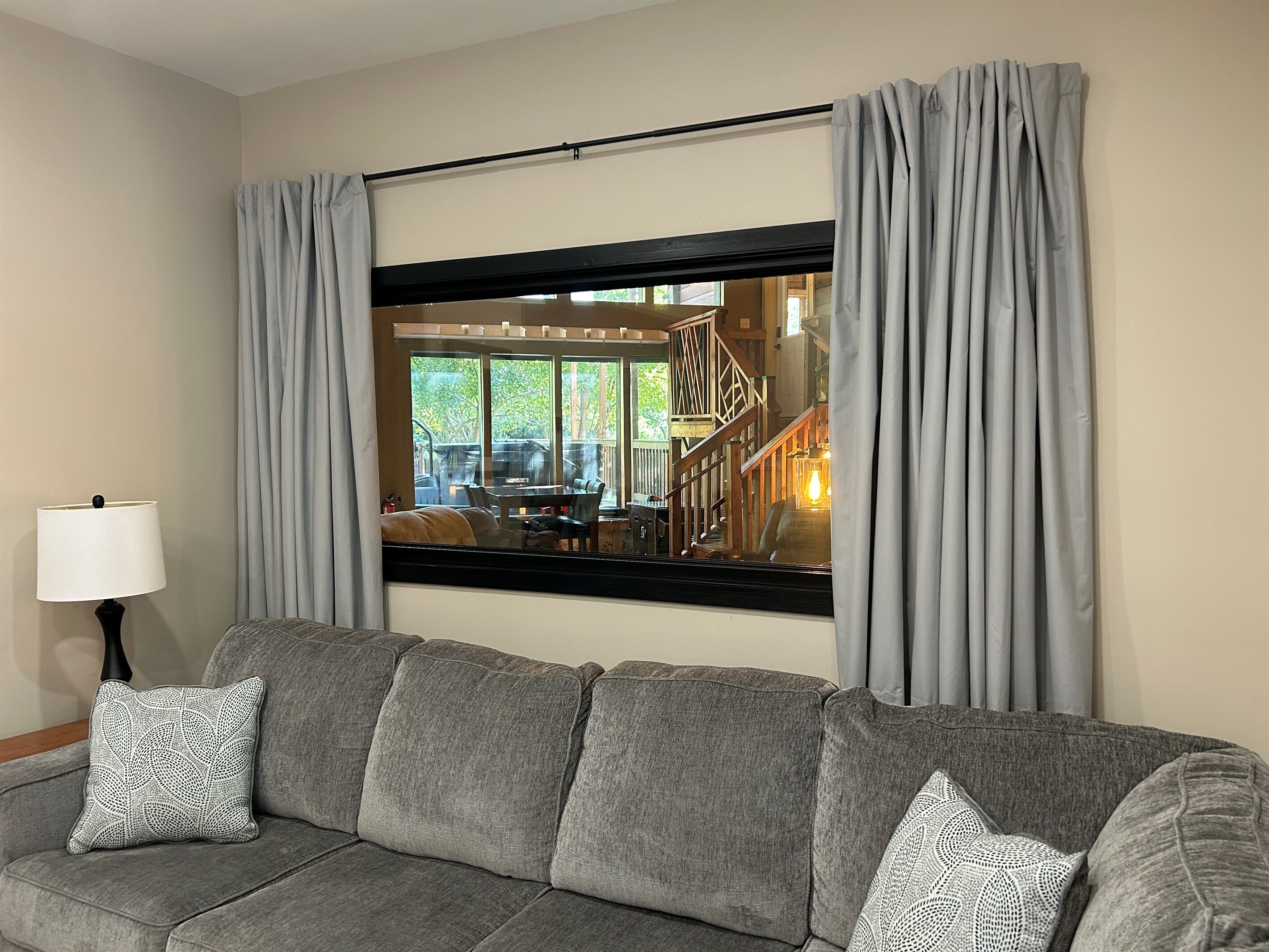 Ein Wohnzimmer mit grauem Sofa, zwei gemusterten Zierkissen, einer schwarzen Stehlampe und einem großen Fenster mit grauen Vorhängen.