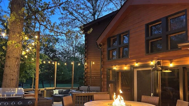 Una terraza de madera con una mesa redonda y sillas, una parrilla y luces colgantes.