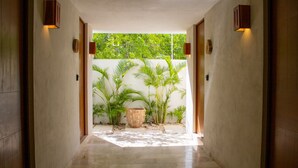 A hallway with a concrete floor leading to a garden area with palm trees and a large decorative pot. - NHOA 107A Lux 2BR Condo Pool access in Aldea Zama (Tulum)