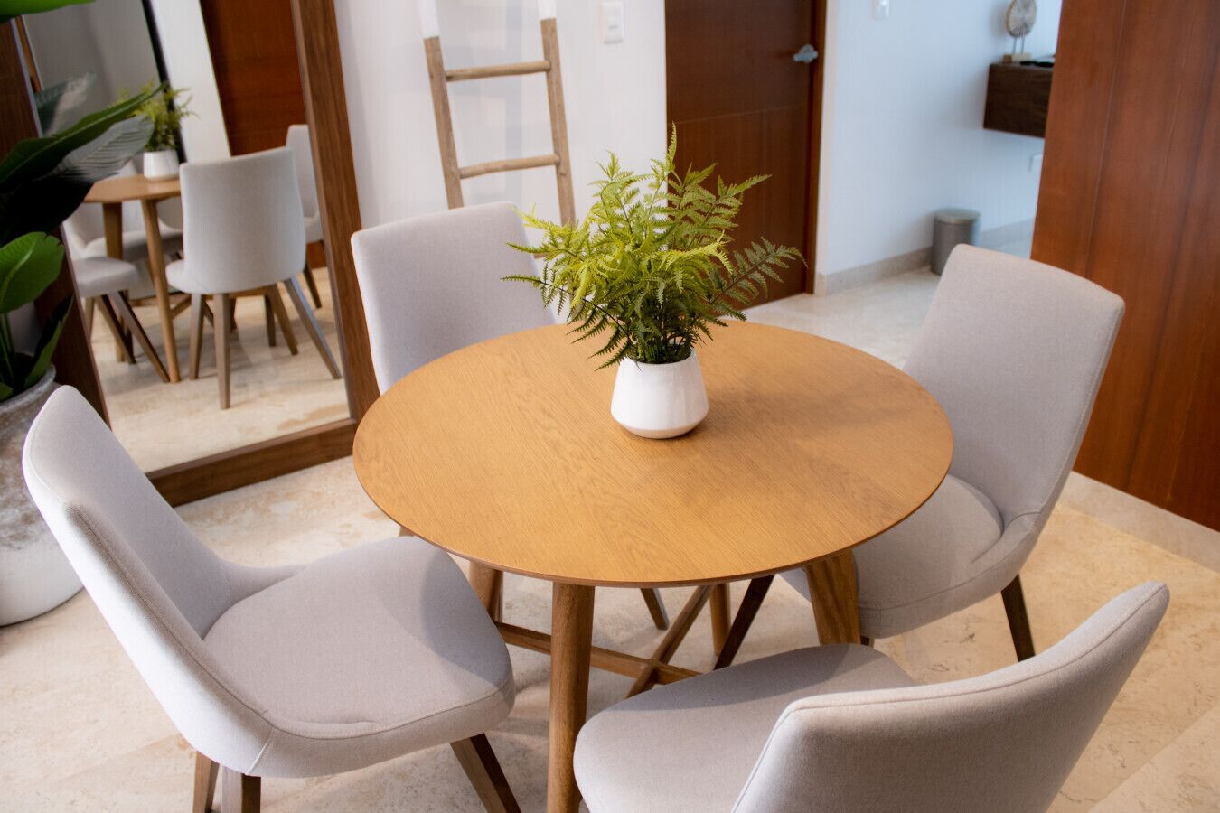 A round wooden table with four grey upholstered chairs, a potted fern, and a wooden ladder in the background.