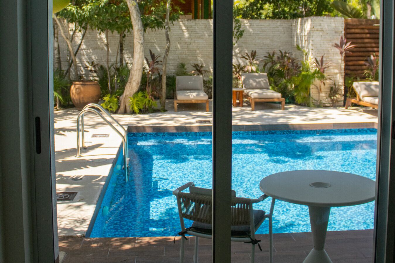 Una piscina con una escalera metálica, rodeada de sillones de descanso y una mesa, con un muro de ladrillo blanco y follaje al fondo.