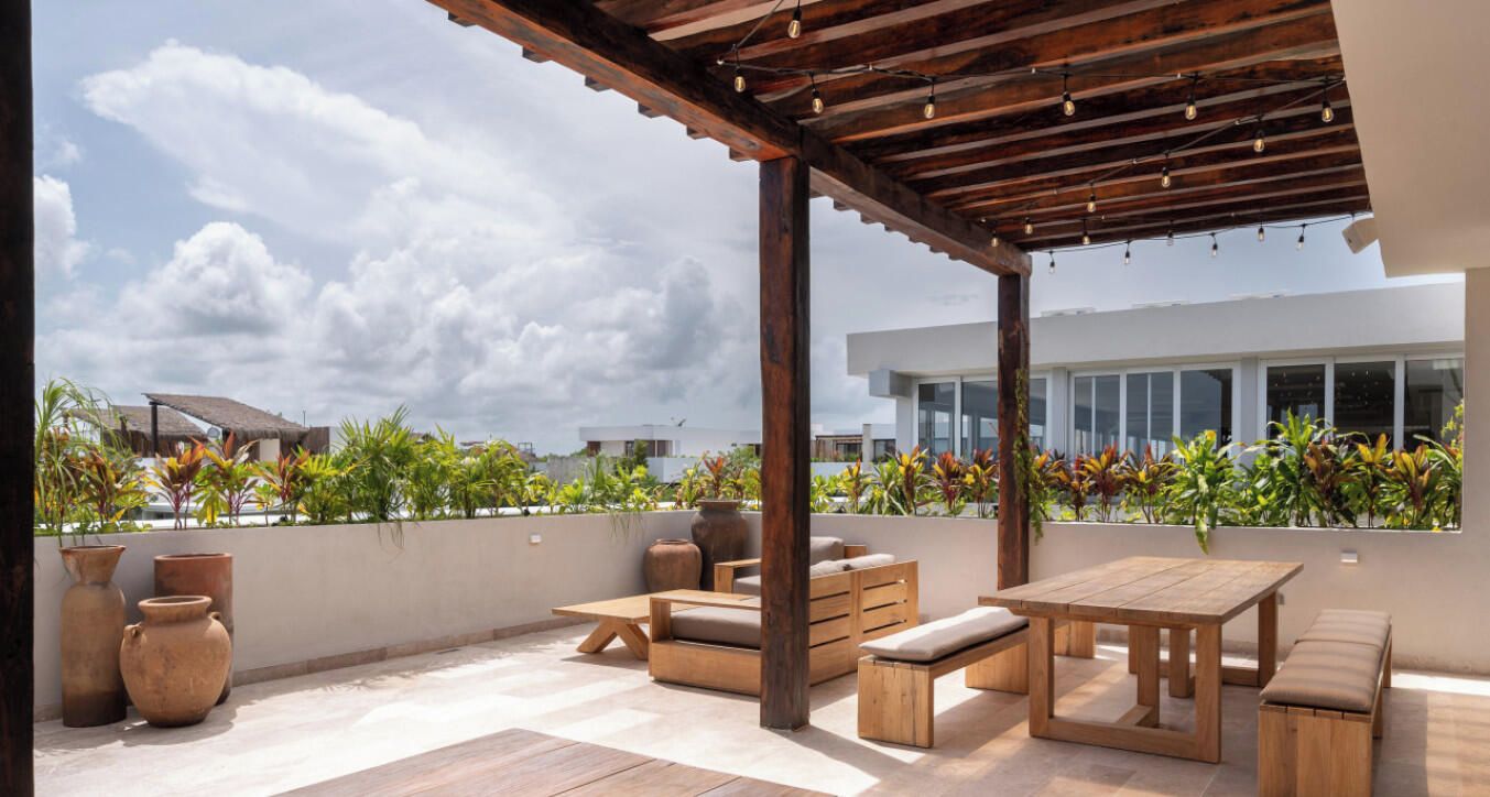 An outdoor patio with wooden furniture, a pergola, and a view of a modern building and greenery.