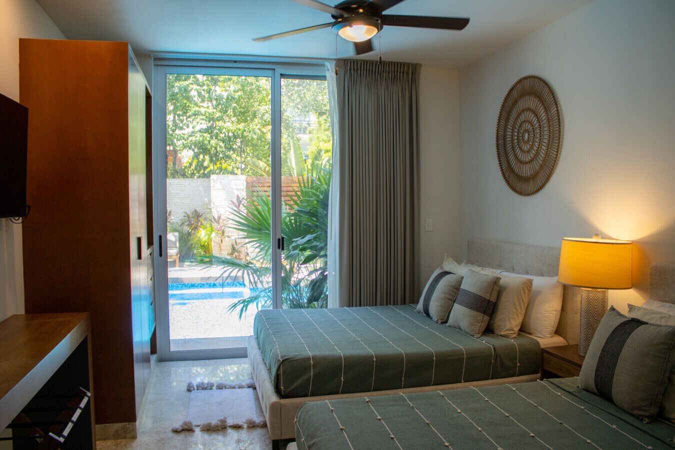 A bedroom with a bed, a nightstand, a lamp, a ceiling fan, and a view of a pool and greenery outside.