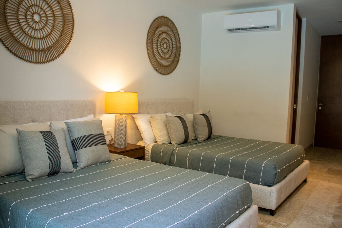 A hotel room with two beds, a nightstand with a lamp, decorative wall art, and an air conditioning unit.