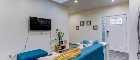 A modern living room with a flat-screen TV mounted on the wall, a blue sofa, a yellow throw blanket, and a small table with a plant and decorative items.