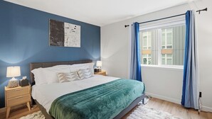 Ein Schlafzimmer mit Bett, Nachttischen, einem Fenster mit Vorhängen und einem Wandbild.
