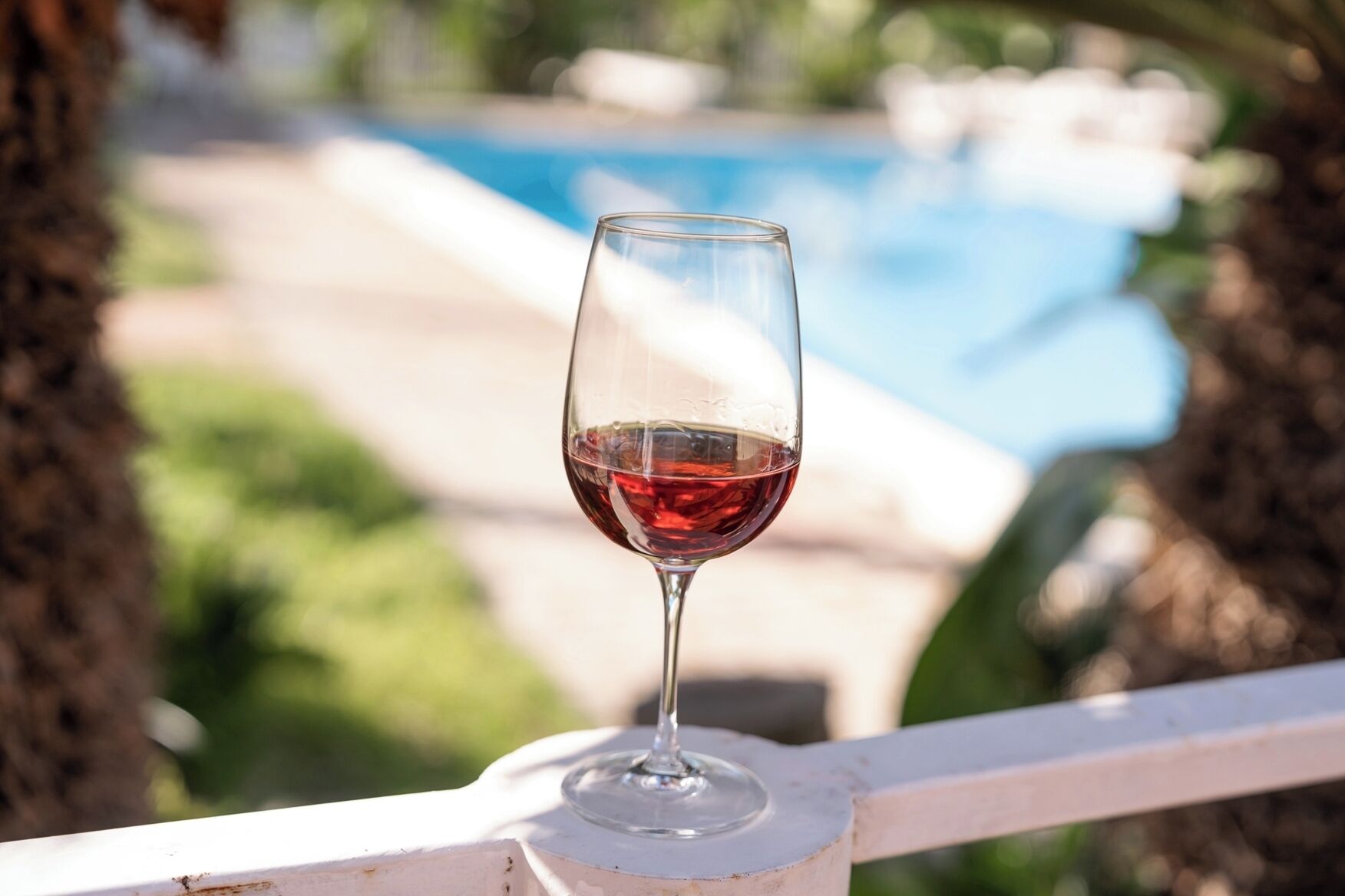 A glass of red wine on a white railing with a pool and greenery in the background.