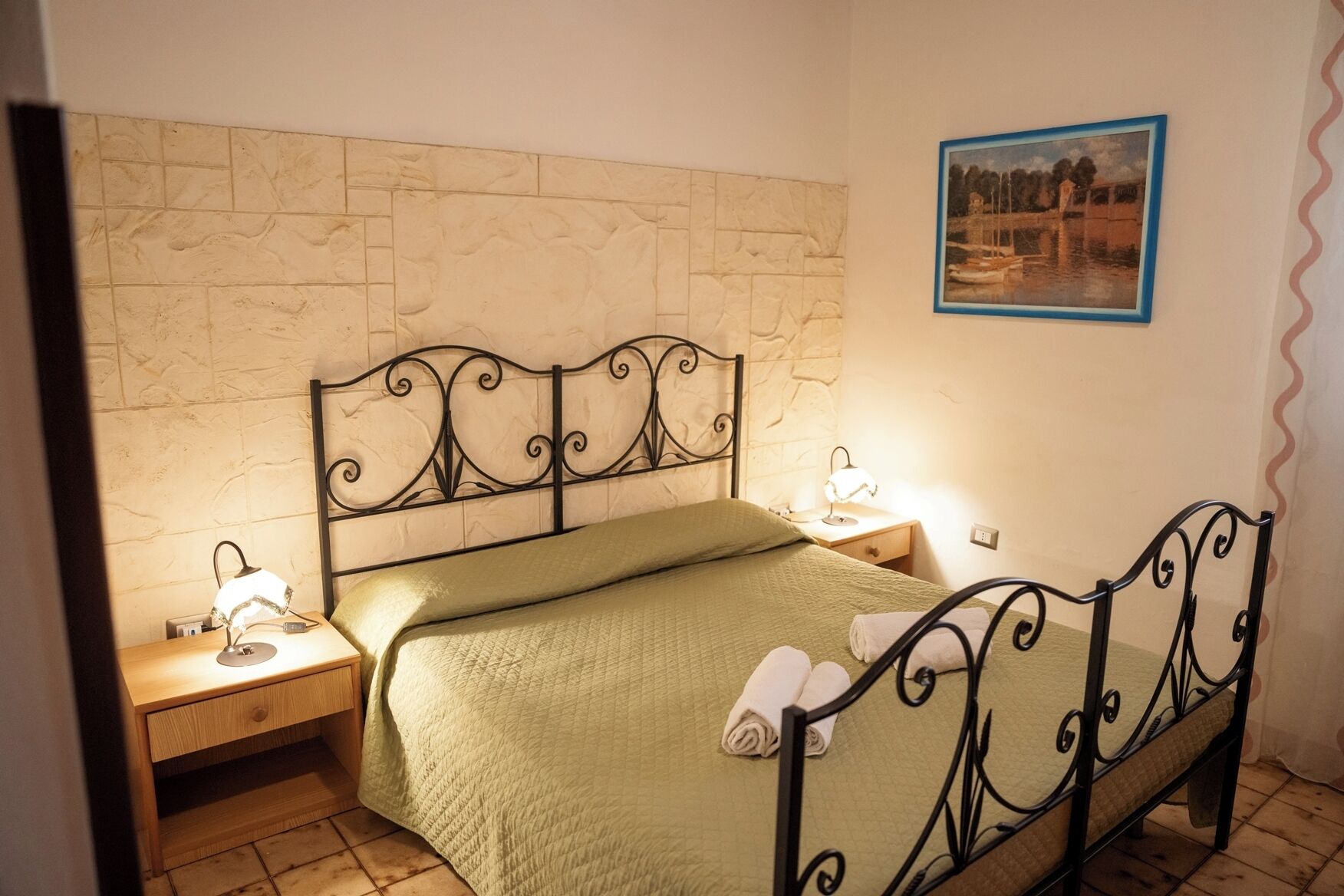 A single bed with a green quilt, two bedside tables with lamps, a stone-patterned wall, and a framed picture on the wall.