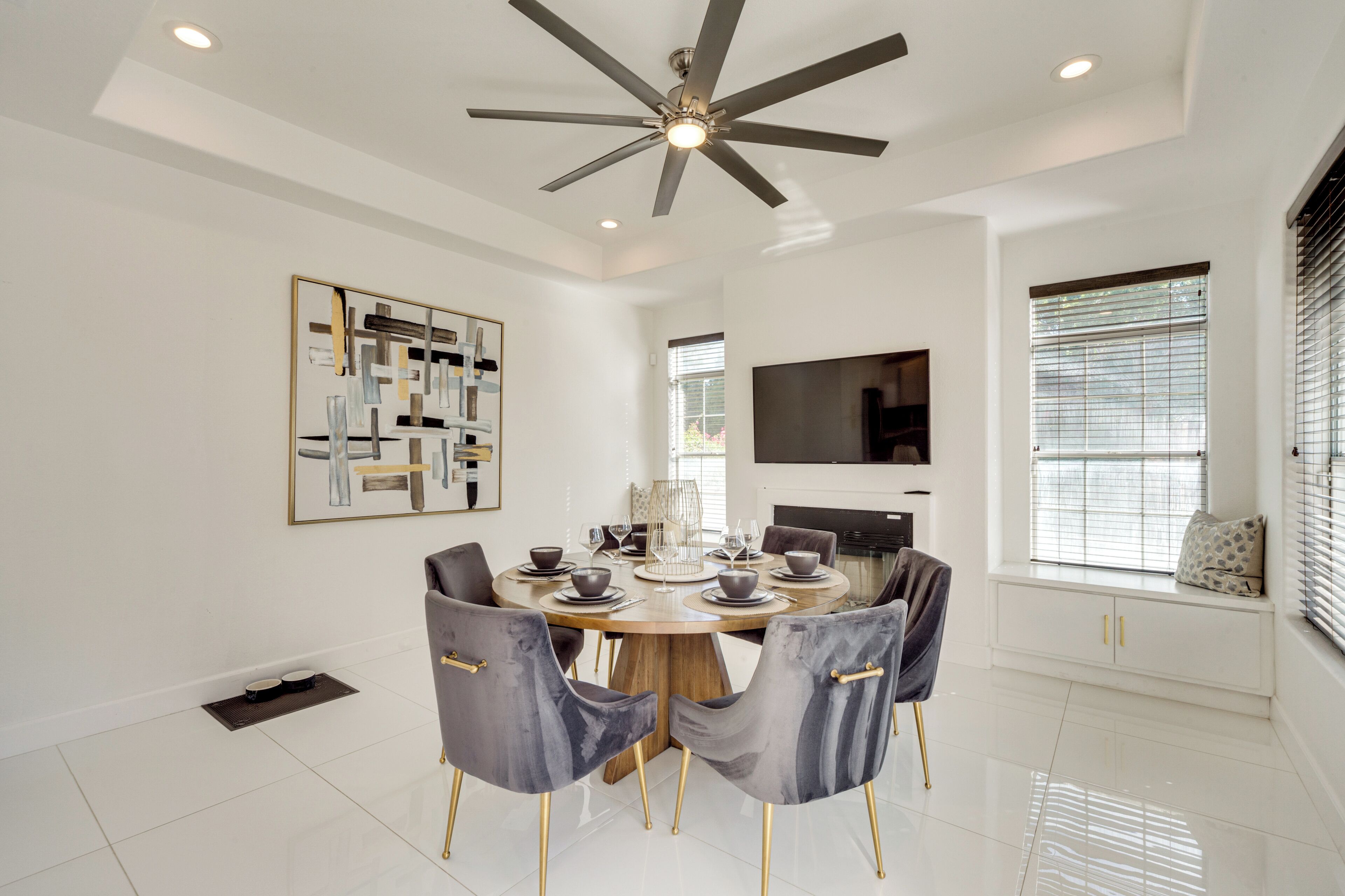 A modern dining room with a round table, grey upholstered chairs, a ceiling fan, and a mounted TV.