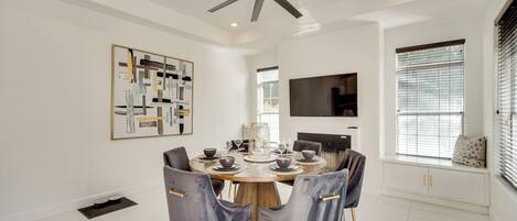 A modern dining room with a round table, grey upholstered chairs, a ceiling fan, and a mounted TV.