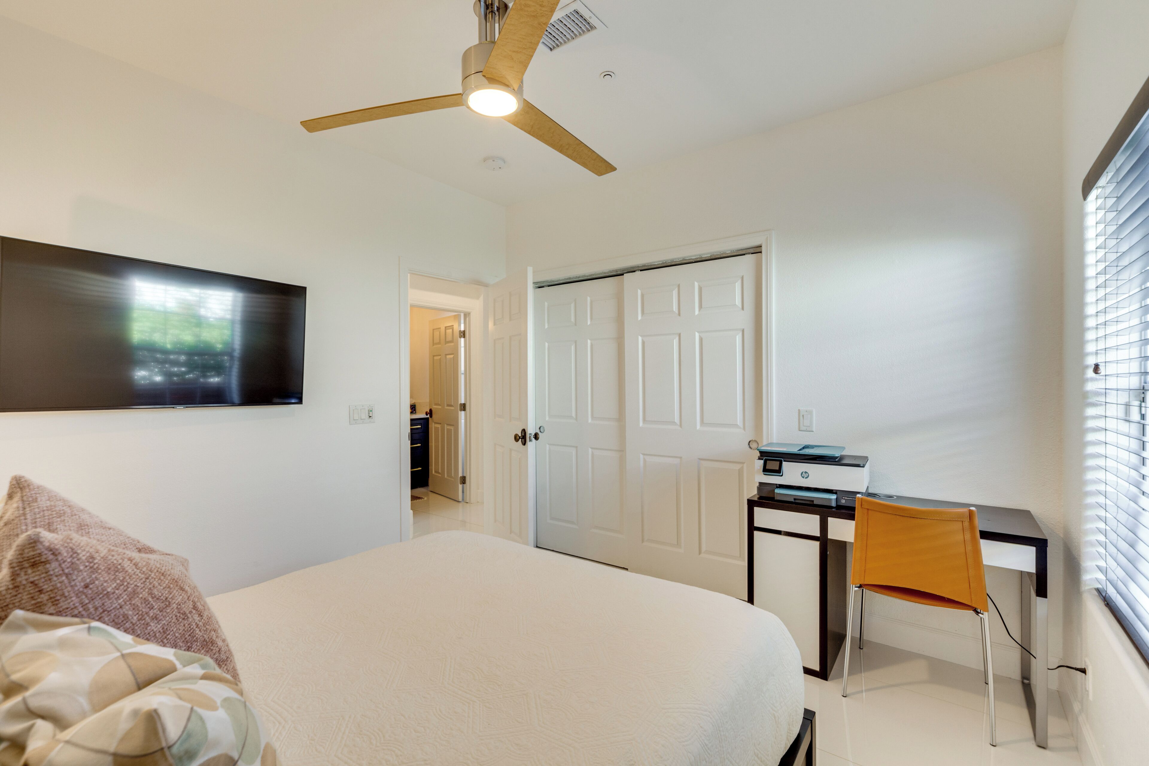 A bedroom with a bed, a desk with a chair, a ceiling fan, and a door leading to another room.