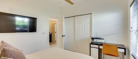 A bedroom with a bed, a desk with a chair, a ceiling fan, and a door leading to another room.