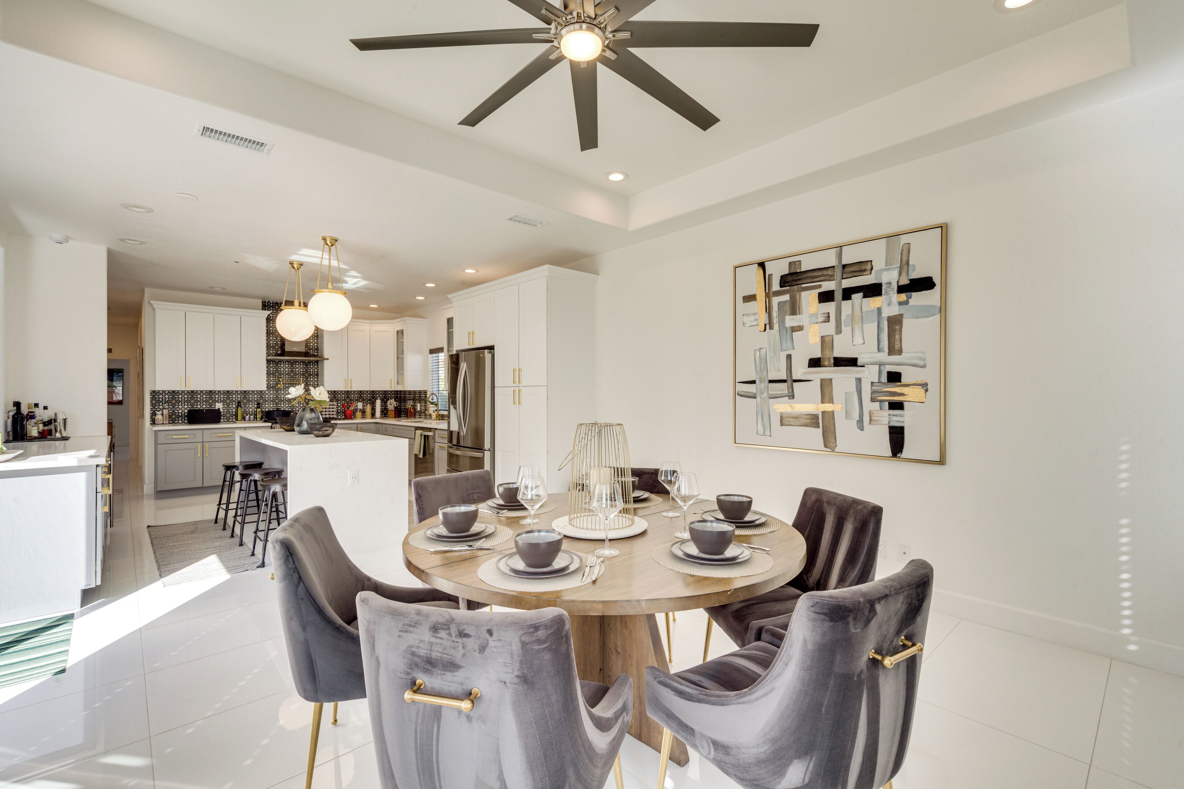 Un espace de salle à manger moderne, avec une table ronde dressée pour six personnes, entourée de chaises grises à revêtement rembourré agrémentées de détails dorés. Un ventilateur de plafond avec un luminaire est visible au-dessus de la table.