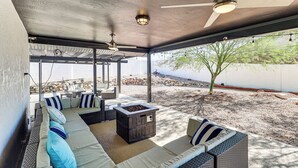 Un patio exterior con una zona de estar cubierta, un pozo de fuego y una pérgola con luces colgantes.