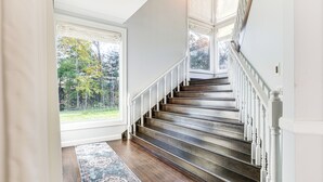 Een trap met witte balustrades en een donker houten afwerking, die uitkomt op een plateau met grote ramen met uitzicht op een groen landschap.