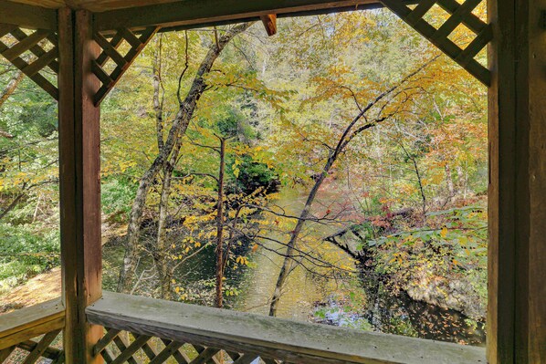 A wooden gazebo with a lattice roof overlooking a forested area with a stream.