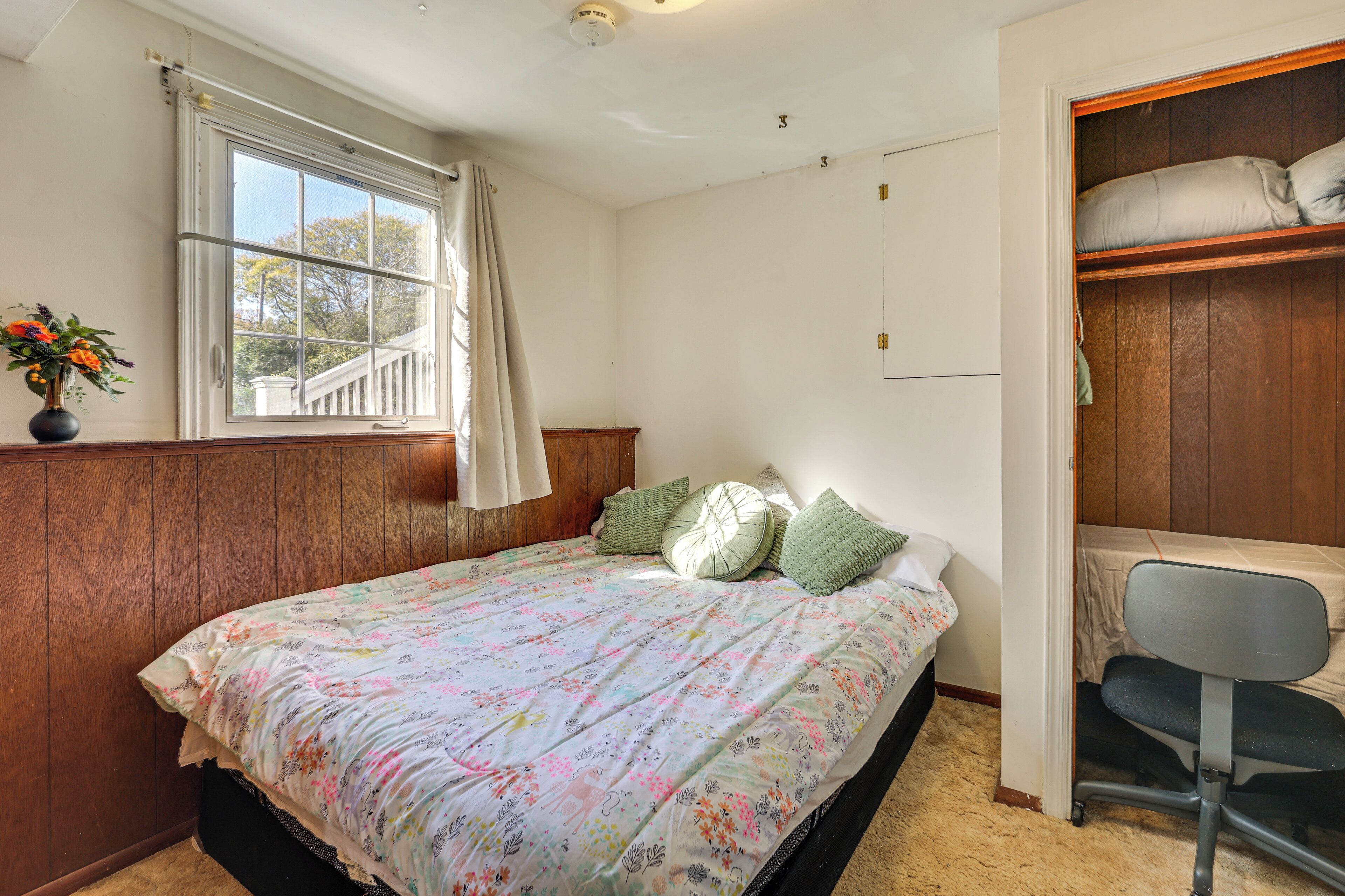 Une chambre à coucher avec un lit, une fenêtre avec des rideaux, un bureau avec une chaise et une armoire avec des portes en bois.