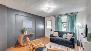 A living room with a dark grey wall, a wooden bench, a black sofa with cushions, a small round table, and a floor lamp. - Little Cove Cottage! Walk to Dtwn Panama City (Panama City)