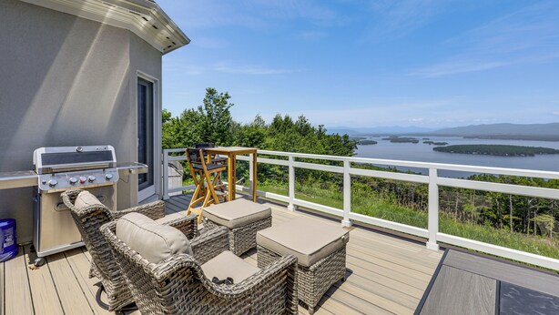 Eine Terrasse mit Gartenmöbeln, einem Grill und Blick auf einen See und Wald.