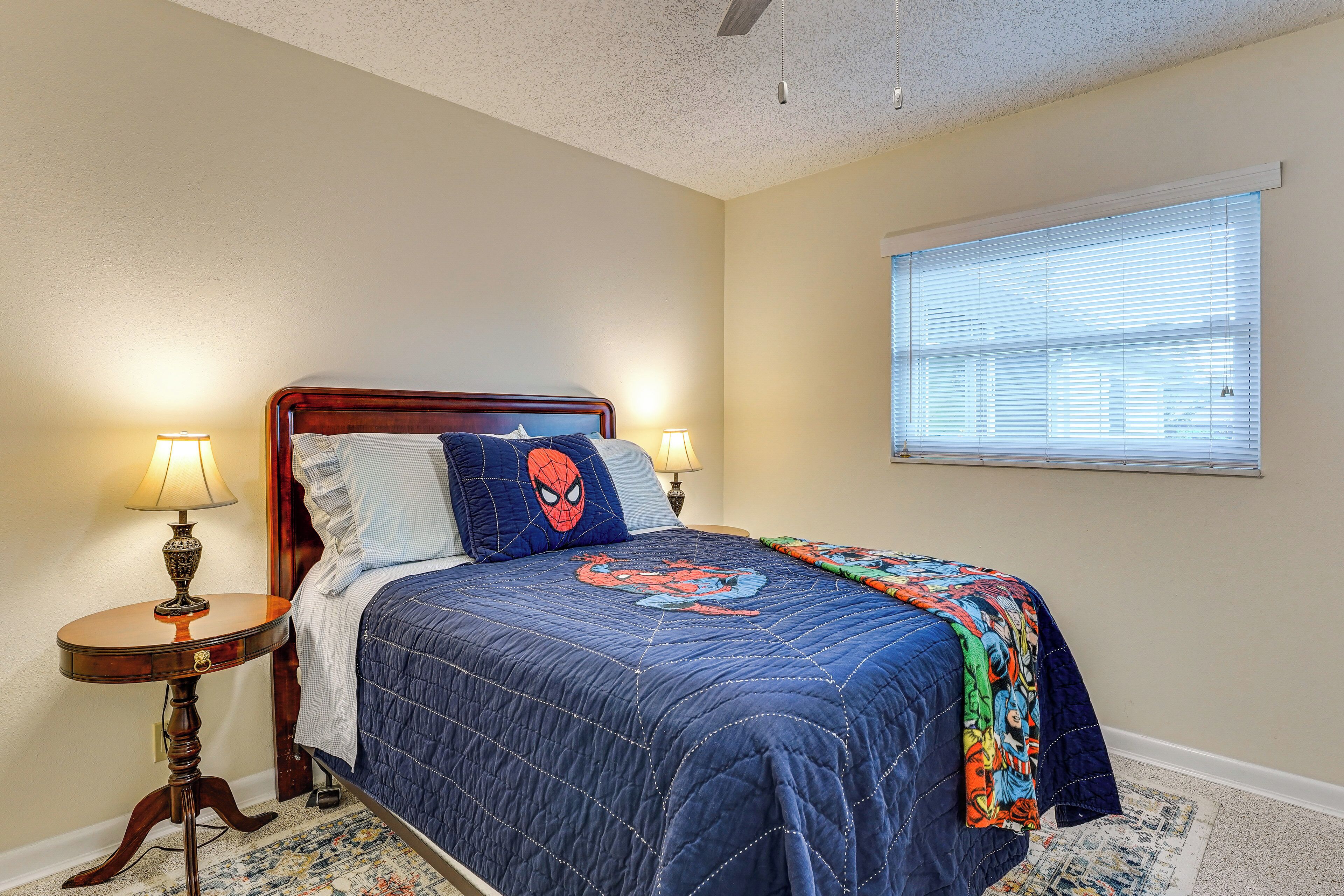 A bedroom with a bed, two bedside lamps, a window with blinds, and a Spiderman-themed bedspread.