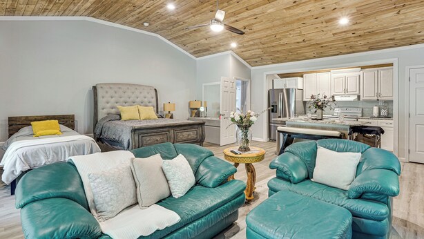 A modern living room with a large teal leather sofa, a matching ottoman, and a tufted headboard bed. A kitchen with white cabinets and stainless steel appliances is visible in the background. - 6 Mi to Clarks Hill Lake: Quiet Couple's Retreat (Appling)