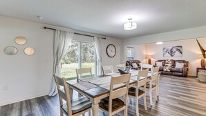 Uma área de refeições com uma mesa e cadeiras de madeira, um relógio na parede e vista para um jardim através de grandes janelas.